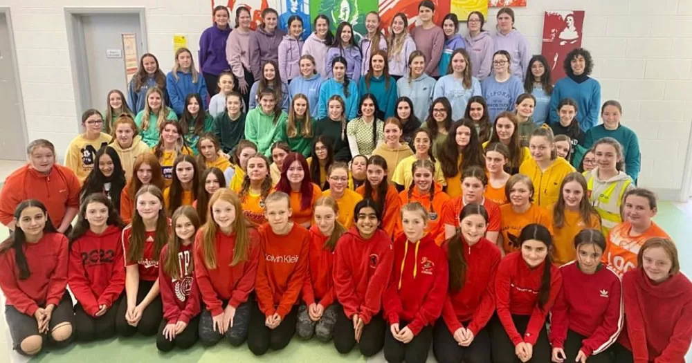 Belong To has launched Stand Up Awareness Week 2023. The image shows a group of approximately 100 students in rows. The pupils in each row are wearing different colour tops to make up a rainbow flag.