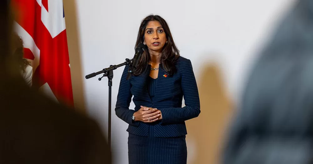 Suella Braverman speaking at a microphone. She is photographed from the thighs up, standing, with her hands folded across her stomach.