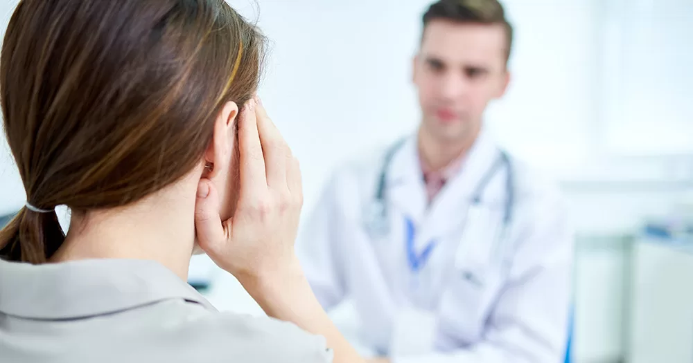 This article is about solving the transgender healthcare crisis in Ireland. The image shows a medical professional consulting with a patient. The back of the patient's head is shown in the left foreground of the photo, with an out of focus doctor facing her.