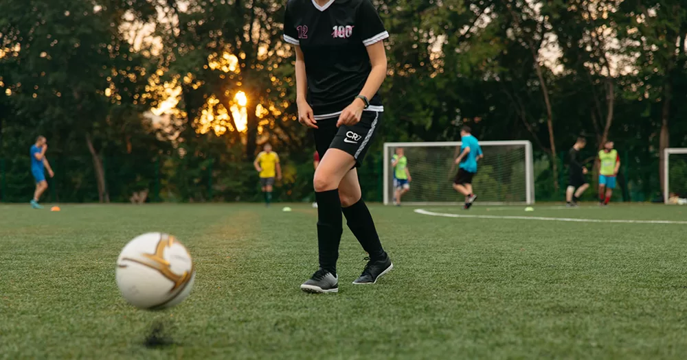 This article is about UN experts encouraging equality for LGBTQ+ athletes. The image shows a person from the shoulders down playing football on an astro-turf pitch. The player wears an all-black kit (jersey, shorts and socks) and a group can be seeing playing football together in the background.