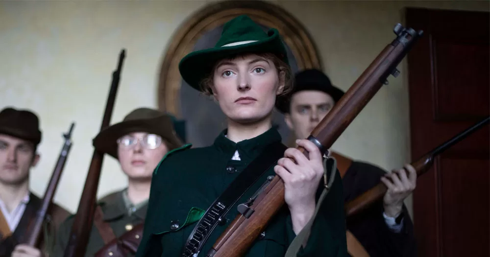 A scene from the new documentary about queer women of the Irish Revolution. The image shows a women standing in front of three other people. They are all wearing clothing from the 1910s. She is wearing an Irish military uniform of emerald green and holding a rifle in her two hands.