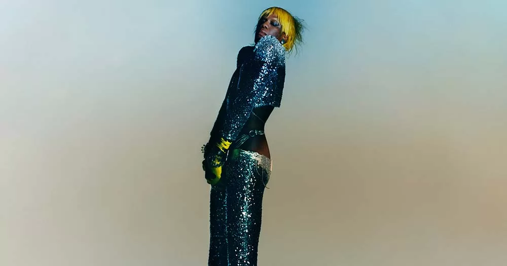 Yves Tumor poses in sequinned bodysuit against pastel background