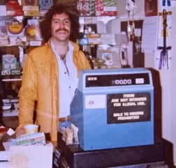 The image shows a photograph of Andy Cramer when he was young, standing behind a cashier desk in one of his stores.