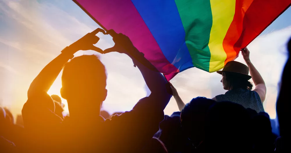 This article is about LGBTQ+ milestones from 2023. The image shows someone holding a rainbow flag above their head, while another holds their arms up with their hands making the shape of a love heart.