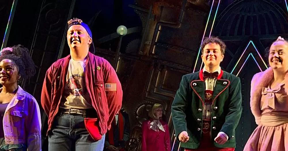 The image shows two male characters from Charlie and the Chocolate Factory the Musical dancing on stage. The person on the left is wearing a wine coloured bomber jacket over a white tshirt with a backwards blue baseball cap. The person on the left has dark curly hair and is wearing green traditional German lederhosen.