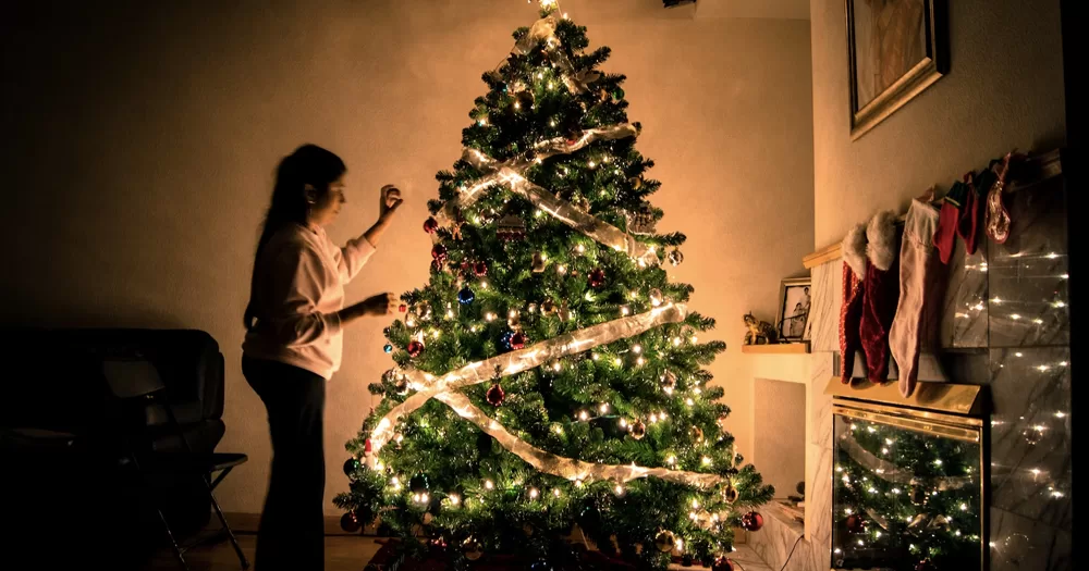 This article is about Christmas songs. In the image, a person decorating a Christmas tree with a lot of lights.