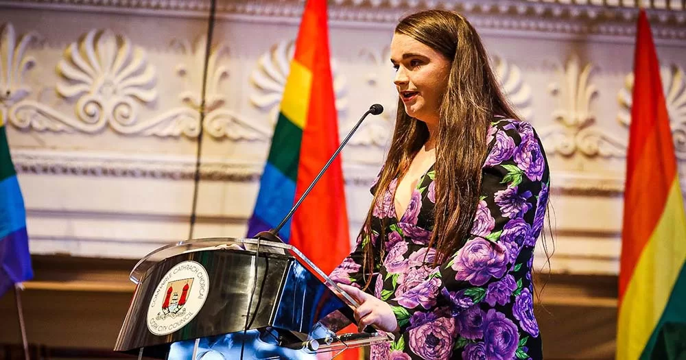 Photo of Saoirse Mackin speaking at podium surrounded by Pride flags at Cork