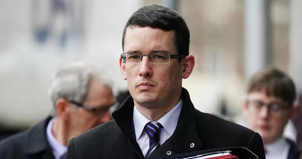 An image of Enoch Burke, who lost a High Court challenge to remove a "promoter of transgenderism" from his appeals panel. He is photographed from the shoulders up, wearing a black coat, white shirt, tie and glasses. He has a serious expression.