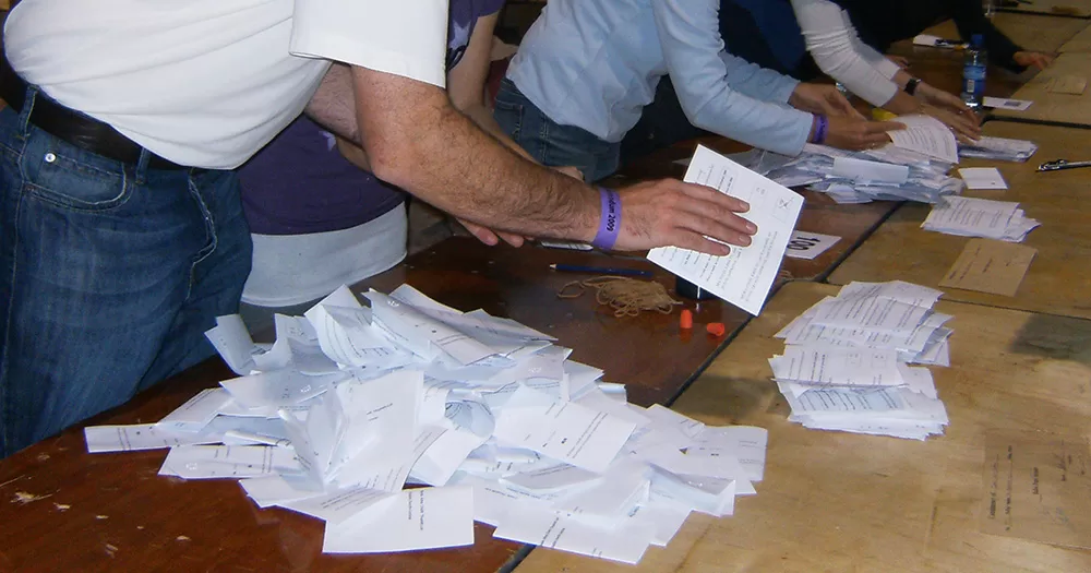 Photo of vote counting at Irish referendums. The images shows slips of paper on a wooden table as a several people leaning over counting.