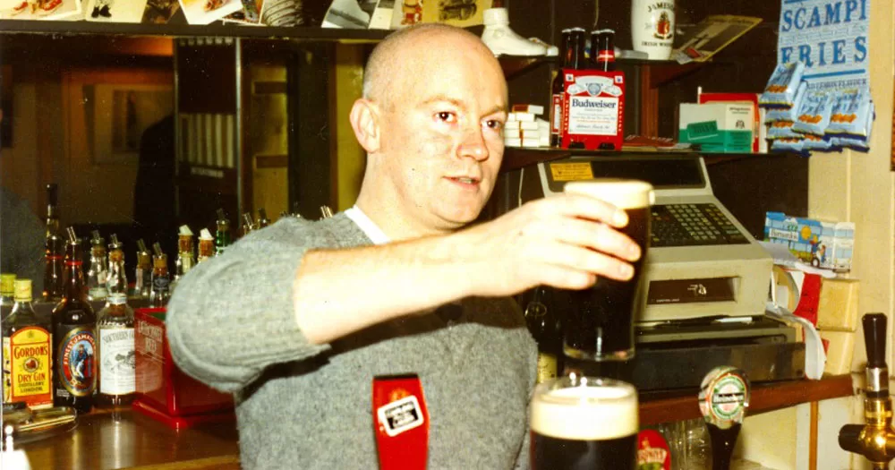 The image shows a photograph used in the new LOAFERS documentary. It shows a man serving a pint of stout across a bar. The photograph is taken on a film camera and dates to the 1908s so the colour is saturated and slightly faded. The man is wearing a grey jumper and has his arm raised to shoulder height as he hands a pint forward. On the counter in front of him is another pint of stout and behind him are bottles of alcohol.