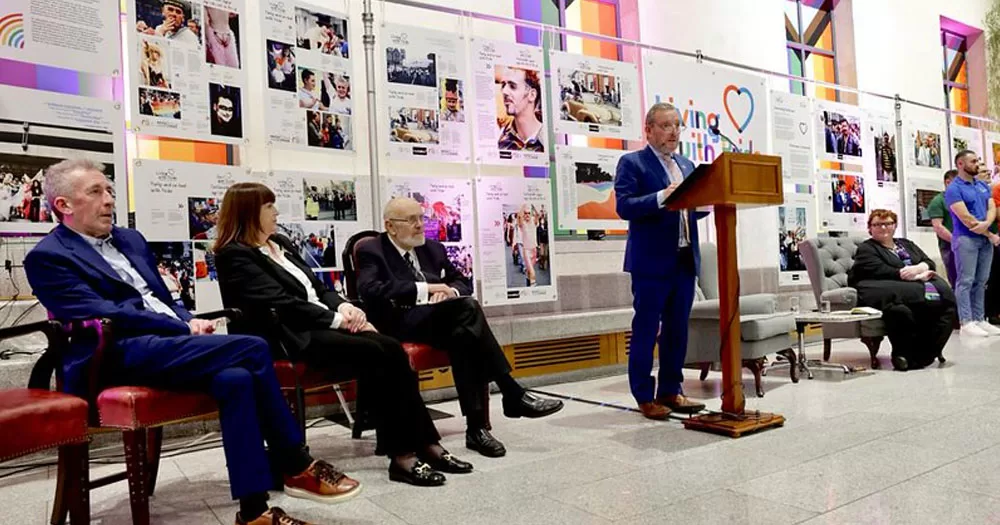 The photo shows three people, two men and one woman, sitting on chairs to the left of a man standing at a podium. In the background, to the right, sits another woman. Behind them is a display of panels from the Living with Pride exhibition.