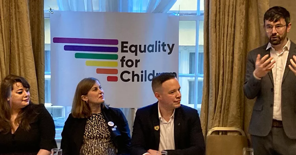 The image shows members of LGBT Ireland, Equality for Children and Irish Gay Dads during a briefing on the amendments to the new surrogacy bill. In the photograph three people are sitting at a table with a sign behind them which reads "Equality for Children" with rainbow coloured stripes. A man wearing a grey suit, glasses and a beard is standing to the right of the image gesticulating with his hands.