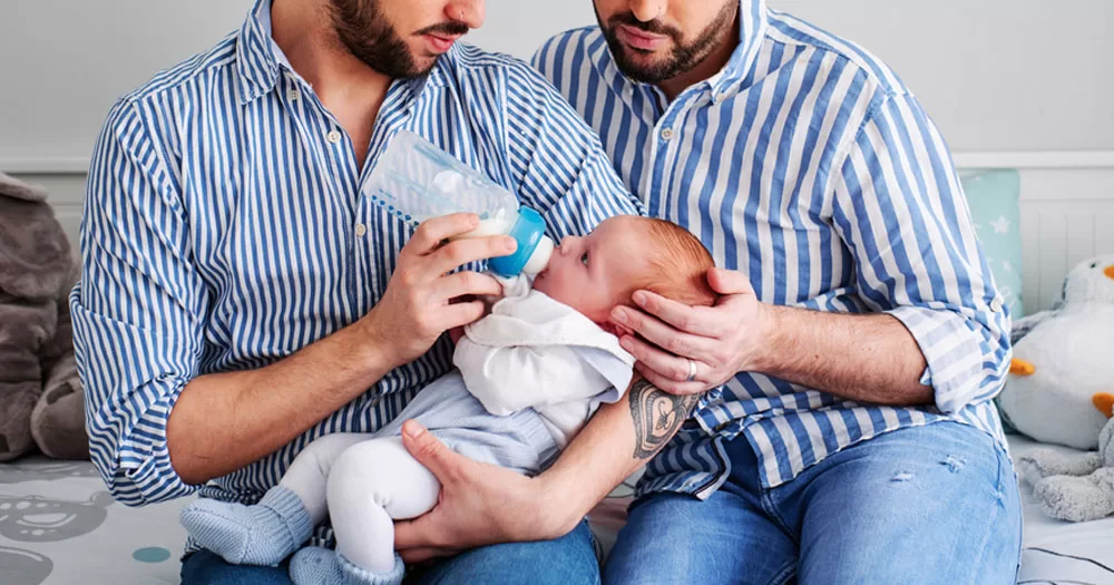 A new bill on surrogacy and assisted human reproduction is to pass cabinet today. The image shows two men holding a baby and feeding it a bottle. They are both wearing blue and white striped shirts and are sitting on a bed with soft toys. Both men have beards and are looking down at the baby. The baby is wearing a light blue jumpsuit and has blue socks on.