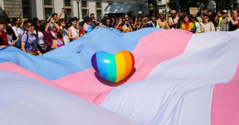 Photograph of a rainbow shaped heart balloon resting in the middle of a giant transgender flag held by a crowd of people, the article is about the RTE transgender healthcare programme