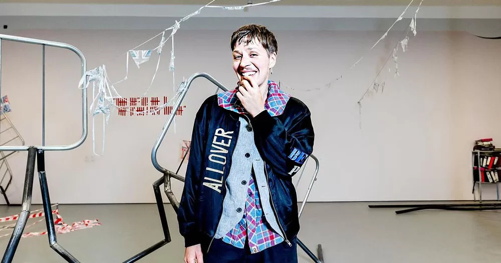 The image shows trans artist Jesse Darling who won the 2023 Turner Prize. In the photo, he is standing in front of metal barriers with fallen bunting of the Union Jack flag. He is wearing a navy sports jacket over a grey waistcoat with a pink and blue checked shirt underneath. He has a slight moustache and is biting his index finger.