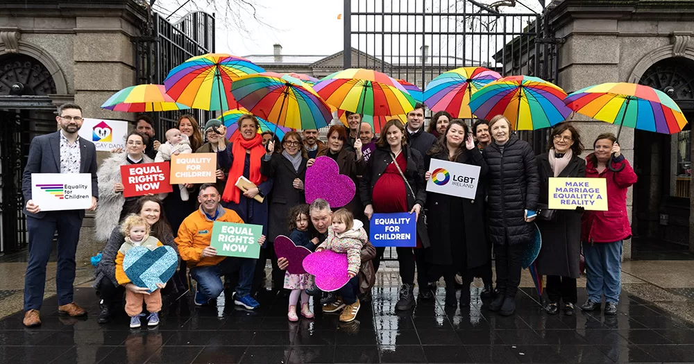 LGBTQ+ families and groups outside the Dáil welcoming a new Bill. The group holds rainbow umbrellas above their heads, and children at the front hold love hearts and signs.