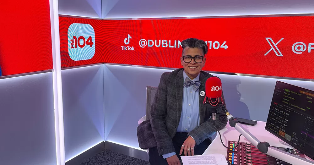 An image of Dil Wickremasinghe, the host of the new FM104 LGBTQ+ show Come In. Dil sits smiling in a radio studio, with red FM104 branding seen in the background.