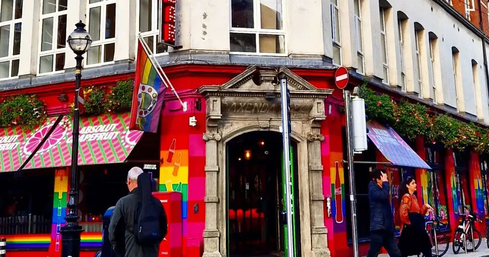 This article is about the history of PantiBar. The image shows a corner building with an ornate old doorway made from grey stone. Around the sides of the building are windows and the walls are painted red with a band of rainbow colours in the middle. There is a flag hanging above the door. It has vertical rainbow stripes and the letter P in the middle.