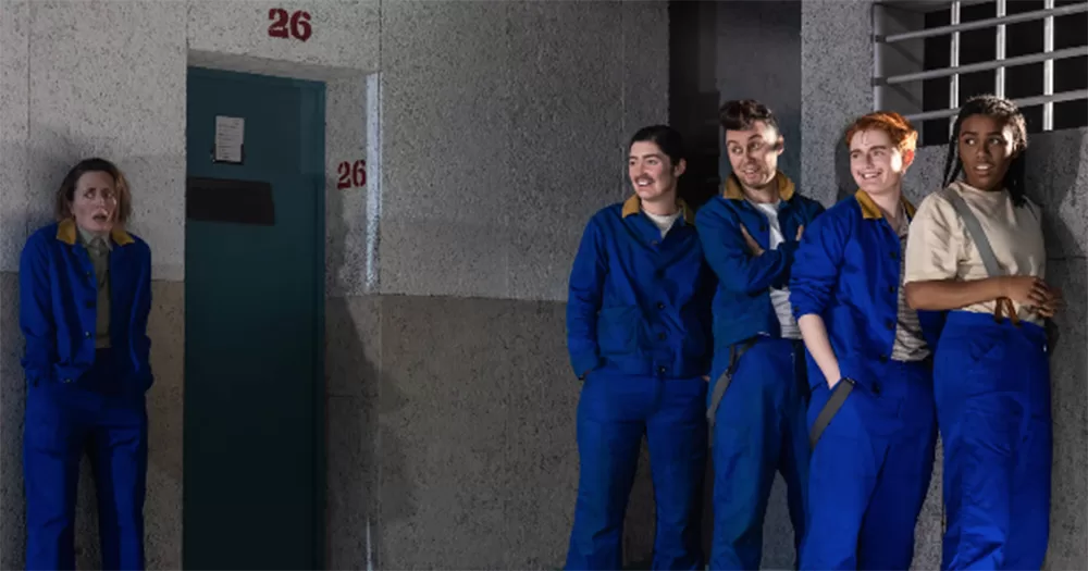 Production image from The Quare Fellow. It shows a group of prisoners, played by women and non-binary actors, wearing blue jumpsuits. Four on the right are smiling, while one on the left looks shocked.