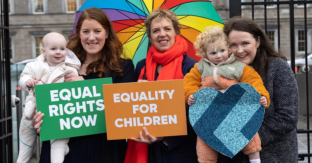 This article is about a new bill proposing amendments to ensure legal equality for children born to same-sex female parents. The image shows Ivana Bacik in the middle, with two women holding young children on either side of her. Ivana holds a rainbow umbrella above her head, and a sign reading "EQUALITY FOR CHILDREN".