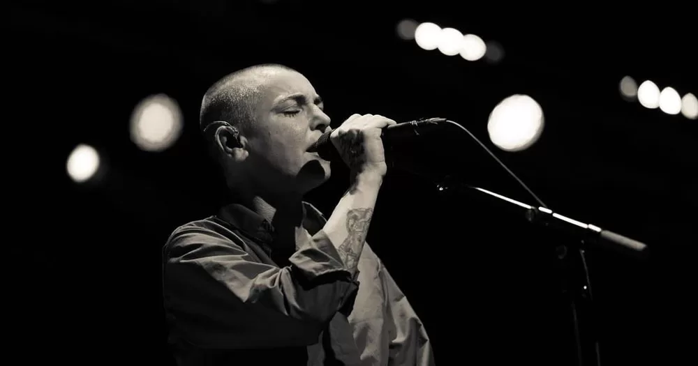 The image shows a black and white photograph of Sinéad O'Connor who is the focus of a new documentary. In the photograph, she is holding a microphone to her mouth and has her eyes closed. Her head is fully shaved and she is wearing a loose shirt with the sleeves rolled up.