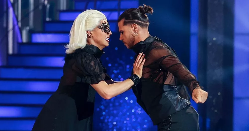 Drag performer Blu Hydrangea and pro partner Simone Arena performing on Dancing with the Stars, facing each other, both wearing black on a blue background.