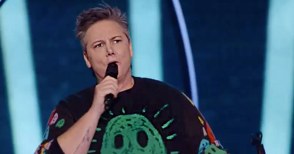 The image shows genderqueer comedian Hannah Gadsby performing on stage. They have show cropped spiky hair and are holding a microphone to their mouth.