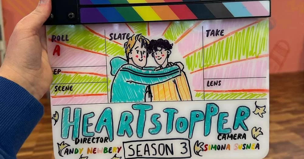Photo of a hand holding the season three Heartstopper clapboard with adorable illustration of Nick and Charlie.