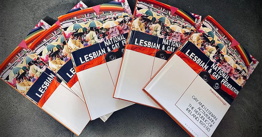 The image shows copies of Gay and Lesbian Activism in the Republic of Ireland which can be won in our History Month competition. In the photo, four copies of the book are fanned out on a table. The lower half of the cover is white with navy and red writing. The upper half shows a photo of people on a float wearing rainbow colours and holding umbrellas.