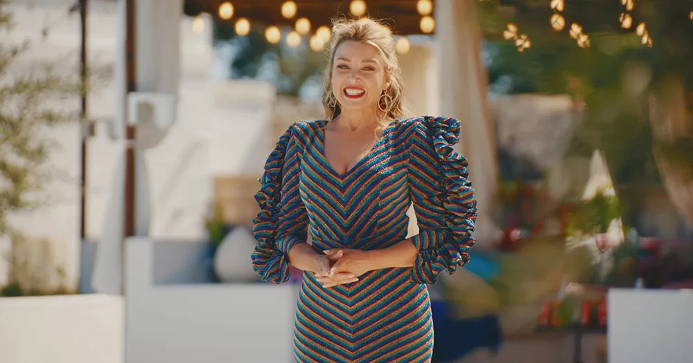 Screenshot of Dannii Minogue hosting I Kissed A Girl. She is photographed from the waist up with a villa in the background. She wears a striped dress and smiles.