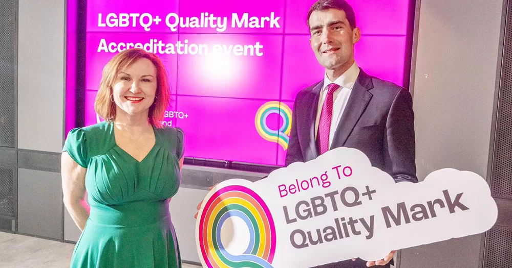 This article is about schools in Ireland receiving their LGBTQ+ quality mark. The image shows Belong To CEO Moninne Griffith smiling with Jack Chambers TD. Jack holds a sign reading "Belong To LGBTQ+ Quality Mark" and a pink screen reading "LGBTQ+ Quality Mark Accreditation event" can be seen in the background.
