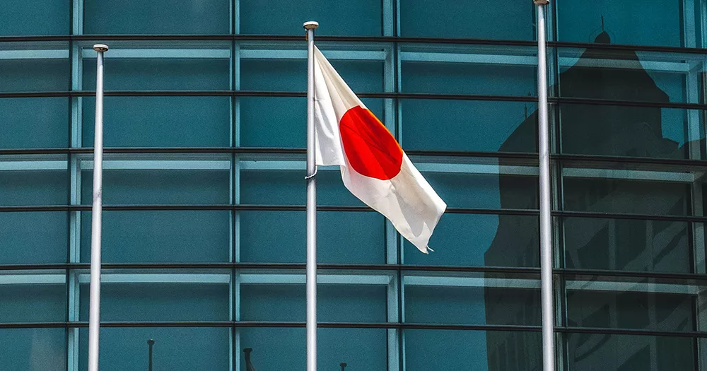 This article is about a trans man in Japan achieving legal gender change without surgical sterilisation. The image shows a Japanese flag flying on a pole, with glass pannels of a building shown in the background.