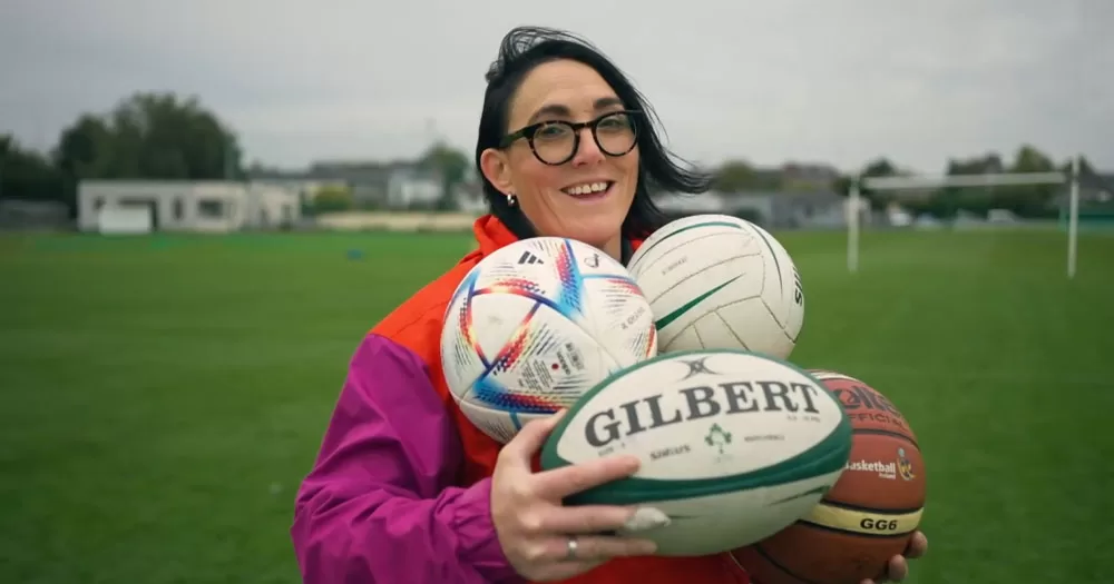 The image is a screenshot from the documentary about Lindsay Peat. In the image she is standing on the side of a sports pitch. She is wearing a pink and red jacket and is holding a soccer ball, a rugby ball, a GAA football and a basketball.