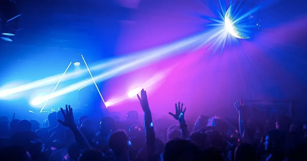 This article is about Mother Daytime Disco Party. In the photo, people dancing in a club with blue and purple lights.