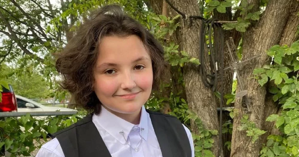 An image of Nex Benedict, a non-binary teenager who died after being allegedly attacked in a school bathroom. They are photographed from the shoulders up wearing a white shirt and grey waistcoat. They look and smile at the camera.