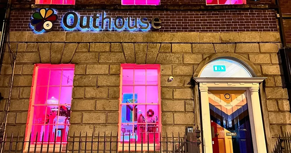 The exterior of Outhouse, which hosts the LGBTQIA+ Alcoholics Anonymous meetings. The image shows the building in the evening, lit up with pink windows and the Outhouse sign illuminated also. The door to the building is painted with a Pride progress flag.