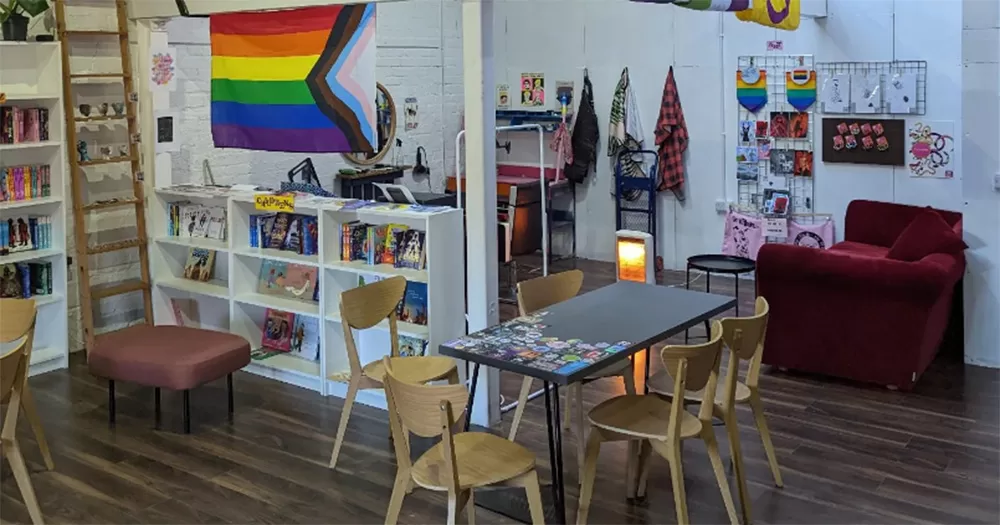 The interior of Paperxclips, Belfast's queer barbers and bookshop. It shows a room with wooden chairs around a table, and a Pride flag on the wall. There are also shelves full of books.