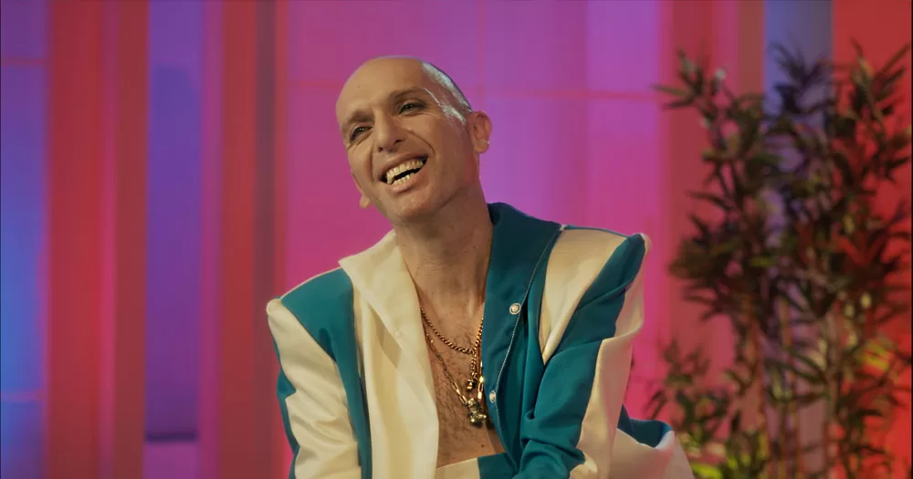 The image shows an outtake from the short film Pregnant with a Drag Queen. In the image, drag queen Veda Lady sits smiling with her head titled to the left. She is wearing a green and lemon blazer open to show a bare chest. She has a shaved head and is wearing gold chains around her neck.