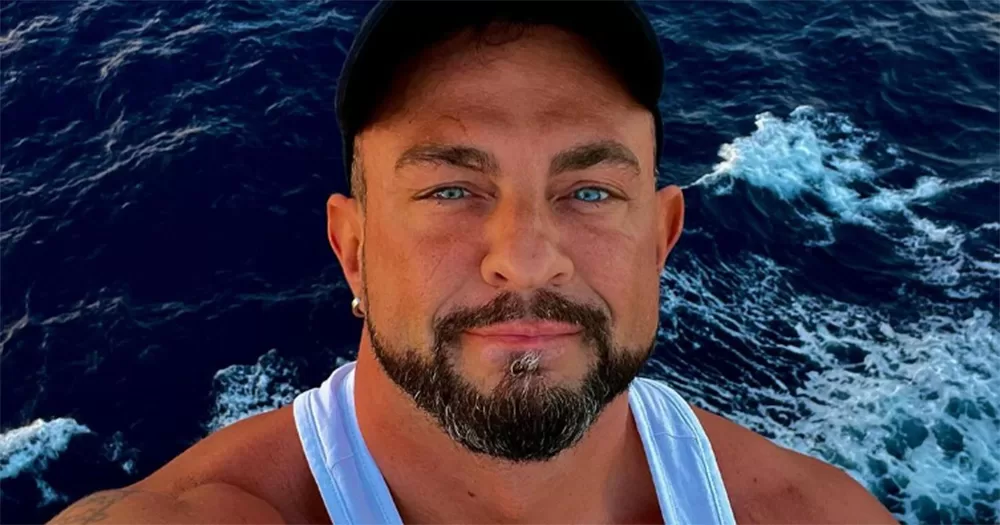 A selfie of Robin Windsor. He smiles at the camera, wearing a black cap and a white vest, with the sea in the background.
