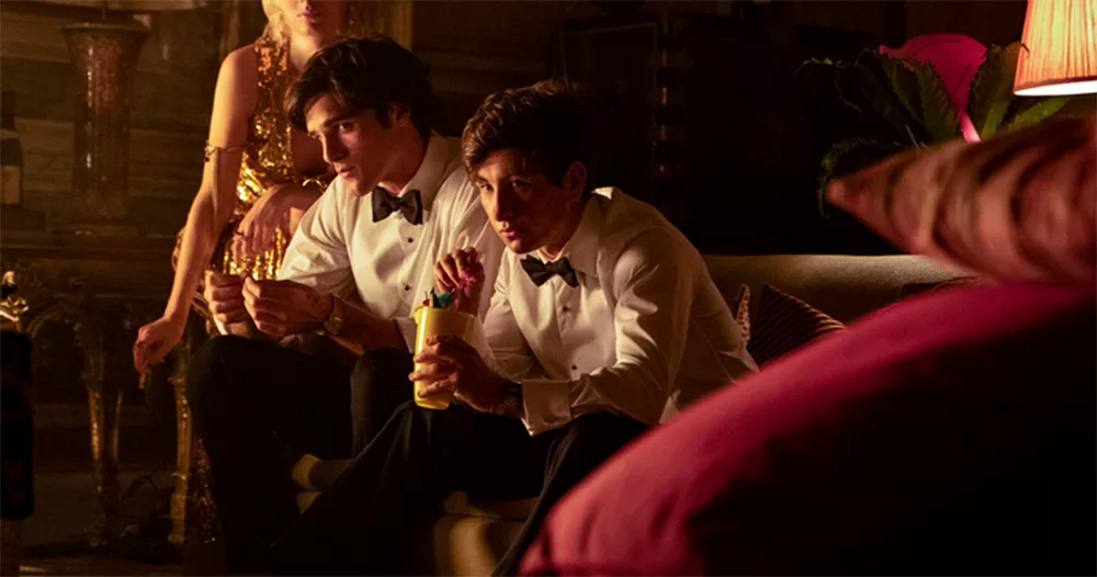 A still from queer film Saltburn. It shows Jacob Elordi and Barry Keoghan as their characters sitting side by side on a couch. They both wear white dress shirts and black dickie bows.