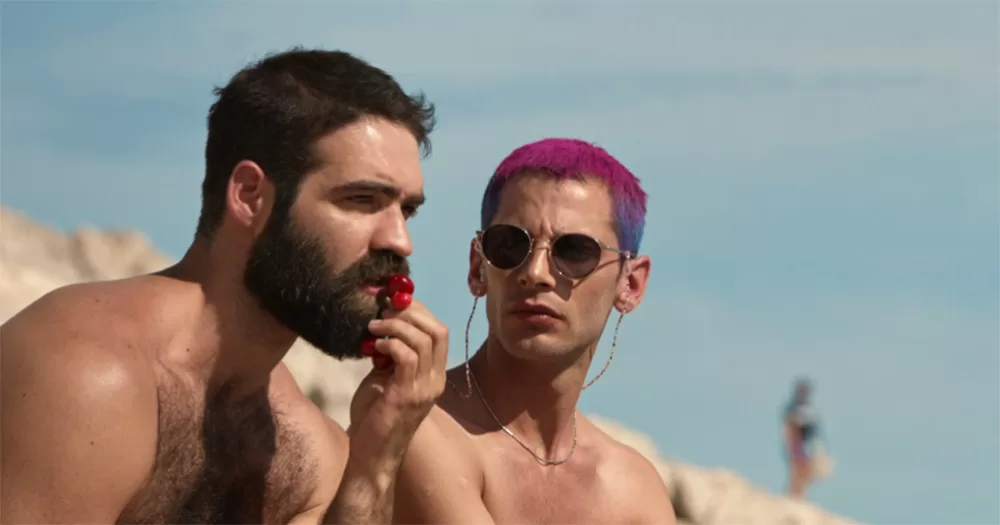 Still image from The Summer with Carmen. It show two Greek men sitting shitless on a beach. The man on the left holds berries up to his mouth as the man on the right, with pink and purple hair, looks at him.