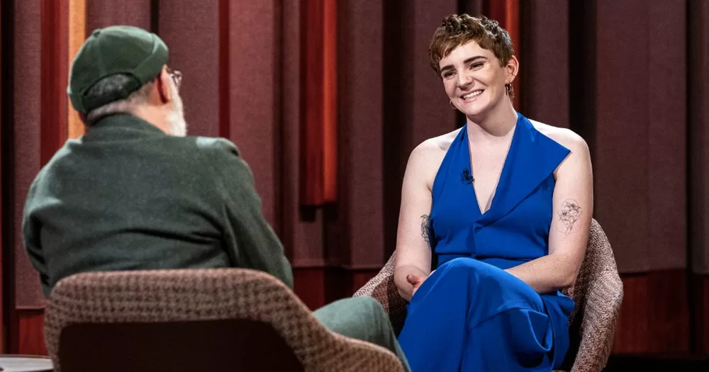 Aoife Commins speaking about HIV on the Tommy Tiernan Show. She is sitting on a chair, smiling, wearing a bright blue suit, while host Tiernan is facing her on an opposite chair.