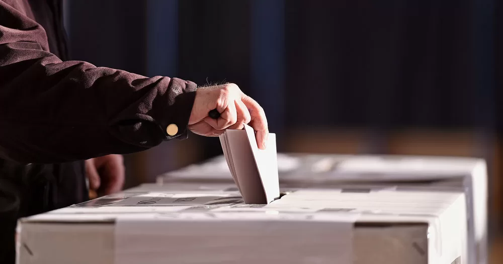 The image shows a hand dropping a vote into a ballot box. People are being urged to vote in the upcoming referendums. The photograph shows an arm wearing a brown shirt pinching a piece of folded paper between the fingers. The paper is halfway into a slit in a ballot box.