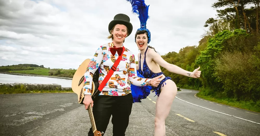 Image of The Wild Geeze, hosts of the No Mother's Day cabaret. On the left is Breda Larkin, wearing a patterned shirt, bow tie and top hat, and right is Miss Lavelle, wearing blue burlesque attire. They smile for the camera, posing in the middle of a country road.