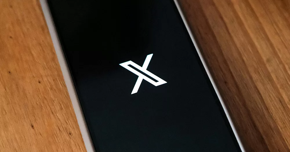This article is about X committing to abiding by Irish hate speech laws. The image shows the X logo on a smartphone. The phone lies on a wooden table.