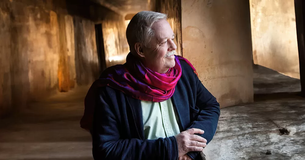The photograph is a portrait of author Armistead Maupin. He is wearing a casual navy jacket over a lime green shirt with a maroon coloured scarf around his neck. He has grey hair and a grey moustache. He is looking to the right, away from the camera.