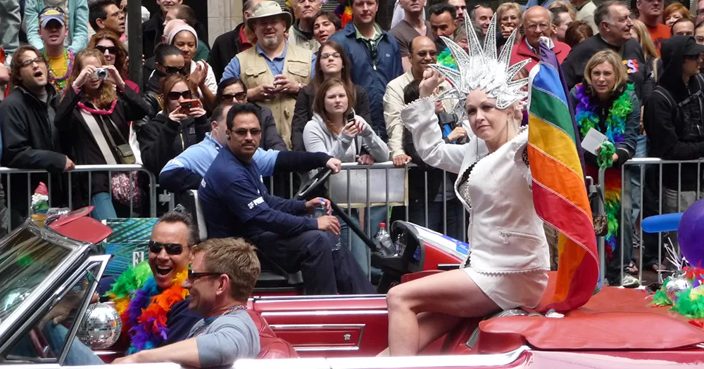 Cyndi Lauper sitting on top of a convertible car at a gay Pride parade. She wears an elaborate silver head piece, and holds a rainbow flag up in the air.