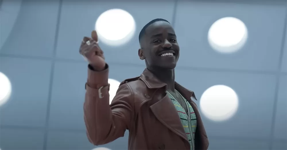 A screenshot of Ncuti Gatwa in the new Doctor Who trailer. He is shown from the waist up, smiling and holding his arm up to click his fingers.