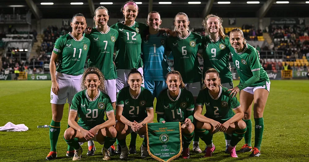 Team photo of the Ireland women's football team who will face England and Sweden in the Aviva Stadium. The players wear their match kits, and smile for a photo on the pitch. They are arranged in two rows, with players at the front crouching and players at the back standing.