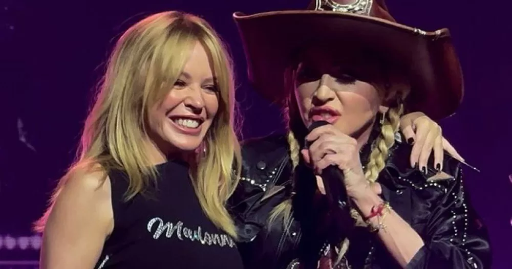Kylie Minogue and Madonna photographed with their arms around each other. Kylie stands on the left smiling, while Madonna stands on the right, singing into a microphone.